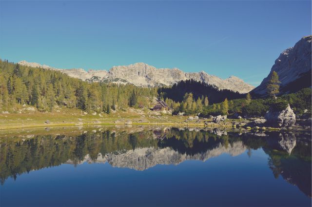 This picturesque scene features a tranquil alpine lake with clear reflections of surrounding trees and mountains. A small wooden cabin is nestled by the shore, adding to the rustic charm. Ideal for use in travel brochures, nature magazines, screensavers, or inspiration for reflecting the beauty of the wilderness.