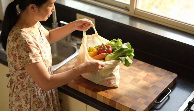 Sunlit kitchen woman unpacking mesh bag of fresh vegetables onto wooden cutting board