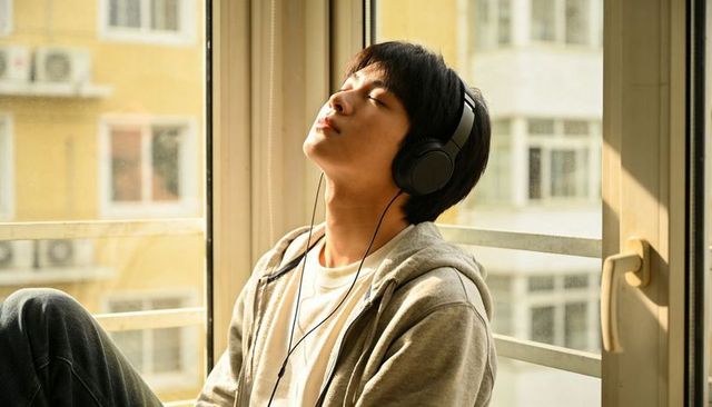 Sunlit young man listening with headphones, relaxing by apartment window with urban view