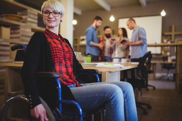 Confident woman in a wheelchair at a creative office with colleagues collaborating in the background. Ideal for use in articles or advertisements about workplace diversity, inclusion, teamwork, and professional environments.