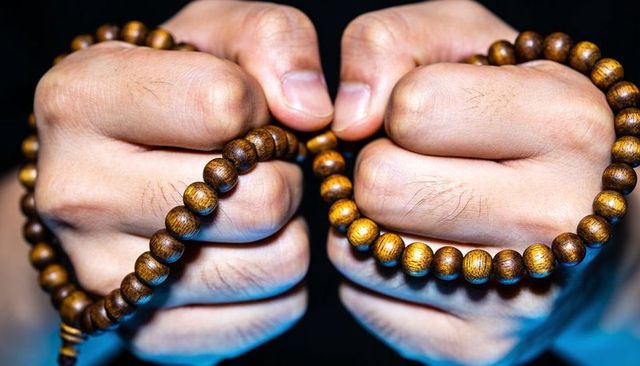 Clasping hands holding wooden prayer beads under dramatic low-key blue lighting, close-up