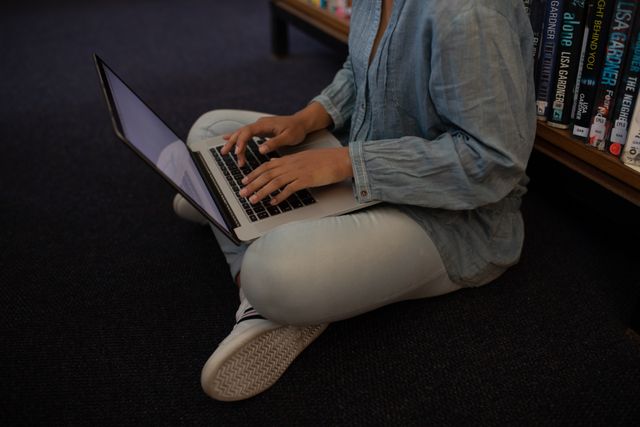 High angle view of low section of a biracial female student wearing a jeans studying in a library sitting on the floor, holding and using computer laptop.