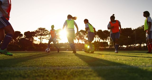 A group of female soccer players are practicing on a field at sunset. They are wearing uniforms with some players in neon vests. The setting sun casts a beautiful golden light enhancing the dynamic and energetic environment. This image can be used to promote sports events, fitness, teamwork, or health lifestyle-related content. Ideal for blogs, sports magazines, training brochures, or advertisements encouraging youth participation in sports.