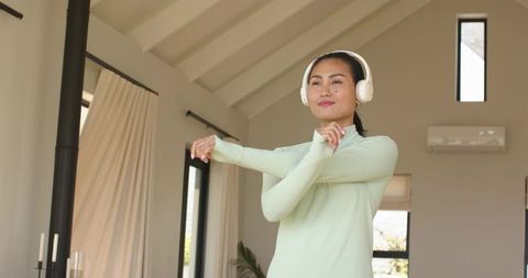 Asian woman stretching at home wearing white headphones and mint green top in sunlit room