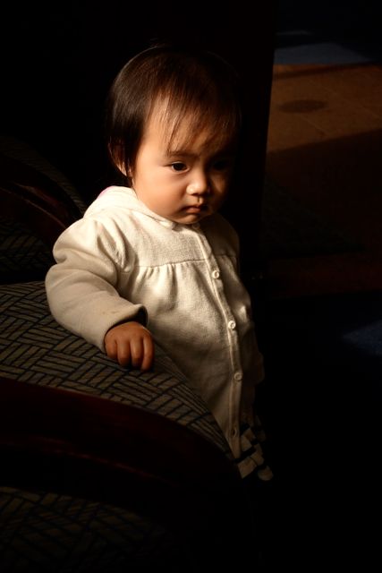 Young male child standing against a sofa in a softly lit room, showcasing a peaceful and introspective moment. Useful for projects related to childhood, innocence, parenting, and home interiors.