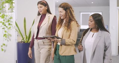 Diverse female team walking office corridor while leader guiding colleagues with tablet
