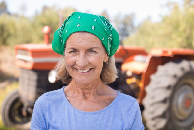 Senior woman standing in front of a tractor in an olive farm, smiling and wearing a green bandana. Ideal for use in agricultural promotions, rural lifestyle blogs, senior living advertisements, and content related to farming and outdoor activities.