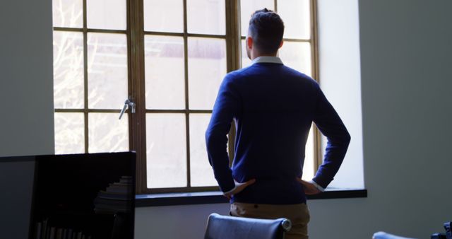 A man in casual office attire stands alone, looking out a large window, hands on hips. This evokes a mood of contemplation and solitude. Ideal for use in articles about workplace dynamics, career planning, introspection, or office-related themes.