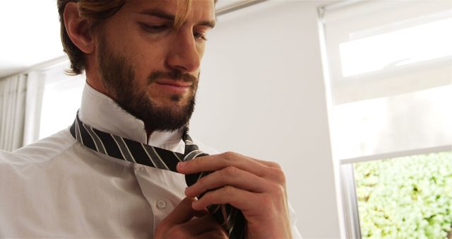 Man tying necktie while wearing white shirt, focusing on preparing for day. Bright room with natural light from windows in background. Suitable for themes of business, getting ready, morning routine, professional attire.