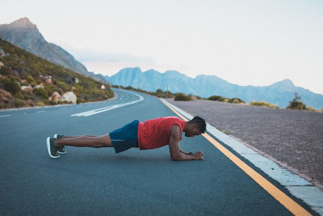 African American man performing a plank exercise on an empty coastal road with mountains in the background. Ideal for promoting outdoor fitness, healthy lifestyle, athletic training, and endurance. Suitable for use in fitness blogs, exercise tutorials, health and wellness websites, and motivational posters.