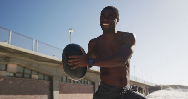 Focused african american man lifting ball, exercising outdoors on beach. fitness, healthy and active lifestyle concept.
