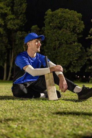 Young male cricketer sitting on grass at night holding cricket bat, contemplative sports portrait