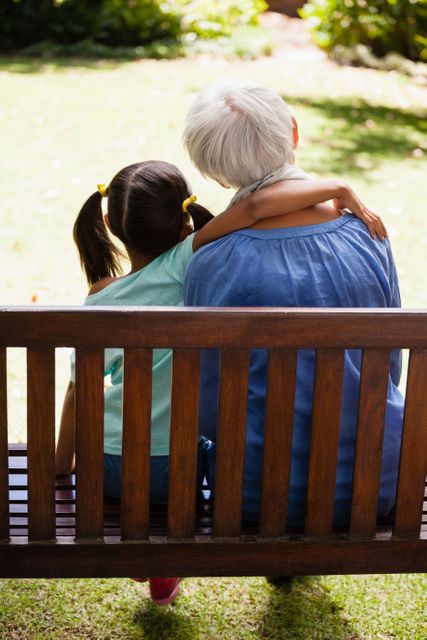 This image depicts a touching moment between a young girl and her grandmother, sitting on a wooden bench in a backyard. The girl has her arm around her grandmother, symbolizing love and bonding. This image can be used for family-oriented content, advertisements for senior living communities, or articles about intergenerational relationships and family values.