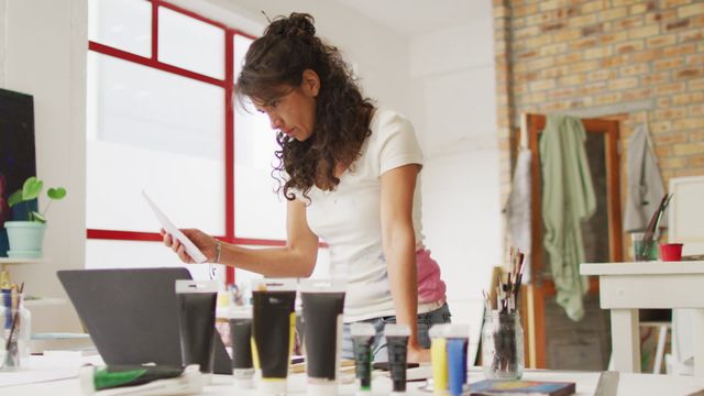 Artist stands at workspace, intensely reviewing her art using laptop for assistance, surrounded by tools, illustrating marriage of creativity and technology. Useful for concepts related to artistic inspiration, the creative process, or technology in art. Ideal for content about artists' workspaces and creativity.