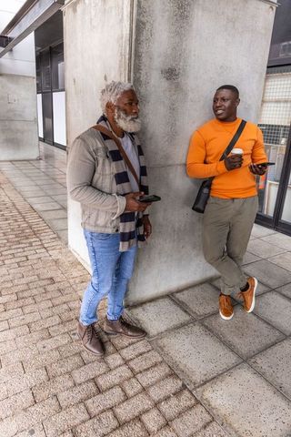 Two african american men leaning and chatting by concrete column holding coffee and phones