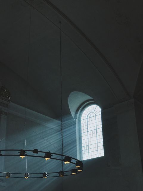 Depicts light beams shining through an arched church window with a chandelier. This serene and spiritual interior showcases the beauty of historical architecture. Would be perfect for use in projects related to faith, spirituality, historical studies, church interior designs, or as a tranquil background for religious ceremonies and events.