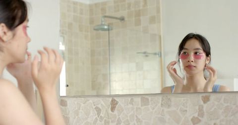 Asian woman using under-eye gel pads at home bathroom mirror