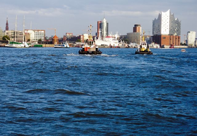 Busy harbor with tugboats sailing across vibrant blue water and contemporary city skyline. Features prominent buildings and Elbphilharmonie. Suitable for articles on modern architecture, transportation, maritime activities or urban tourism.