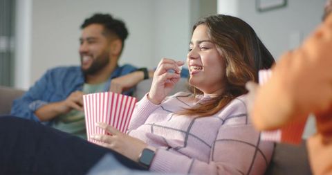 Friends laughing and eating popcorn on couch during cozy movie night at home