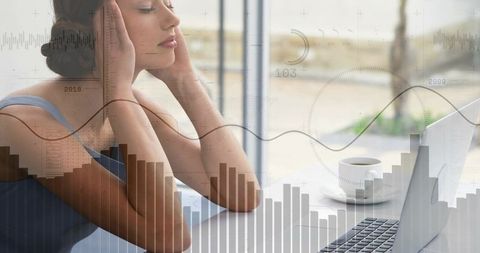 Stressed woman pressing temples while analyzing data on laptop with coffee and graph overlay