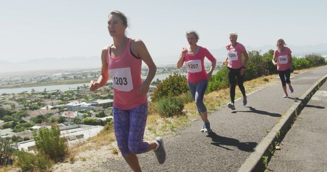 A group of women participating in a marathon run up a hill path with a clear, scenic view in the background. The runners wear pink shirts and number tags. Ideal for use in articles on fitness, outdoor activities, teamwork, women in sports, and promoting an active lifestyle.