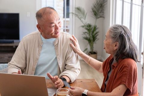 Asian senior couple using laptop and smartphone at sunlit kitchen table