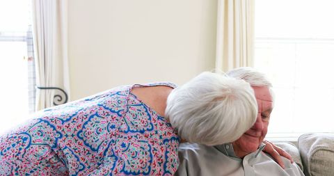 A senior woman is kissing a senior man on the cheek, conveying warmth and affection in a home living room environment. This scenario highlights the tender bonds and love in later life, serving as a representative of healthy aging and strong relationships. Ideal for use in materials related to family values, aging, wellness, and housing for seniors.