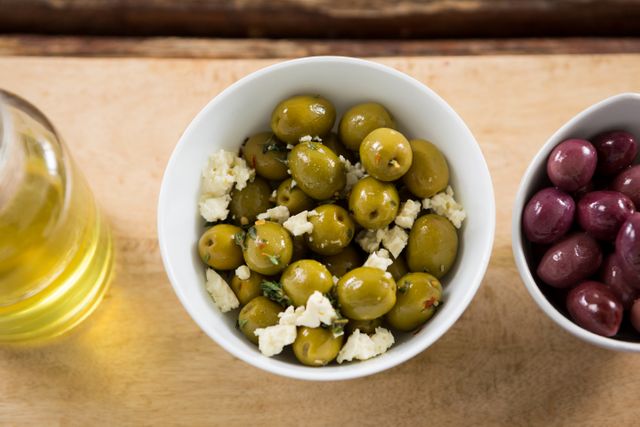 Marinated green olives with feta cheese in a white bowl on a wooden table. Ideal for use in food blogs, recipe websites, or Mediterranean cuisine promotions. Perfect for illustrating healthy snacks, appetizers, or gourmet dishes.