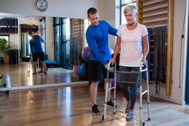 Senior woman receiving assistance from a physiotherapist while using a walking frame in a clinic. Ideal for use in articles or advertisements related to elderly care, physical therapy, rehabilitation, healthcare services, and mobility aids.