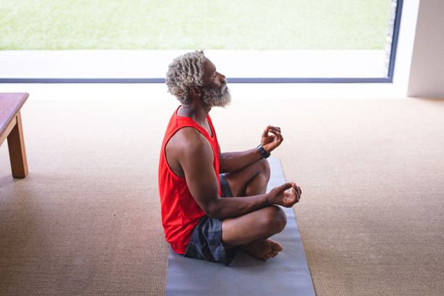 Senior African American man practicing yoga in lotus pose on mat at home. Ideal for promoting mindfulness, wellness, and healthy living. Suitable for articles, blogs, and advertisements focused on fitness, meditation, and active lifestyles for seniors.