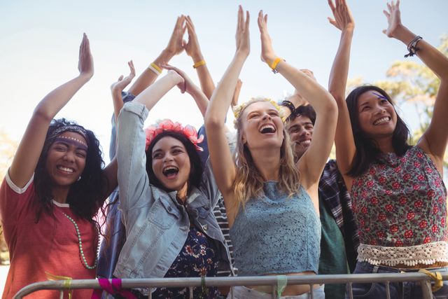 This image captures a group of cheerful female fans enjoying a music festival. They are standing by a railing, raising their hands, and smiling under a clear sky. Perfect for promoting music festivals, outdoor events, summer activities, and youth culture. Ideal for use in advertisements, social media posts, and event promotions.