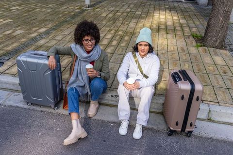 Two women sitting on curb resting with suitcases and holding coffee while waiting