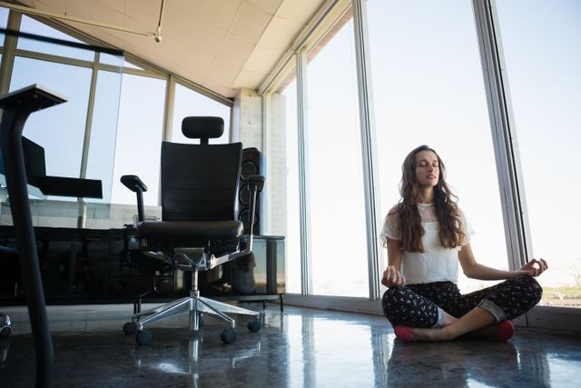 Young businesswoman practicing yoga and meditation on the office floor. Ideal for illustrating concepts of work-life balance, corporate wellness, stress relief, and mindfulness in the workplace. Useful for articles, blogs, and promotional materials related to mental health, professional well-being, and modern office environments.