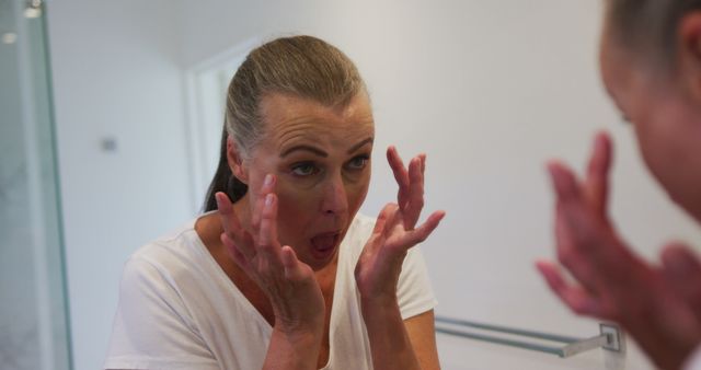 Mature woman in white shirt applying skincare cream to face while looking in a bathroom mirror. Ideal for content focusing on beauty, skincare routines, anti-aging products, and self-care practices. This can be used in advertisements for skincare brands or articles discussing daily beauty routines and health maintenance.