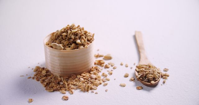 Granola is spilling out of a wooden cup, with a small wooden spoon beside it on a light surface. Excellent for use in advertisements promoting healthy eating, breakfast options, or natural food products. Suitable for use in blogs, articles, or brochures related to nutrition and healthy lifestyle.
