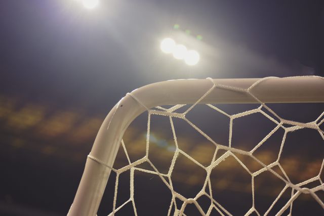 This image captures a close-up view of a soccer goal's net with stadium lights shining brightly in the background during a nighttime event. It can be used for sports event promotions, soccer match advertisements, articles about soccer equipment, or illustrating the competitive nature of night games.