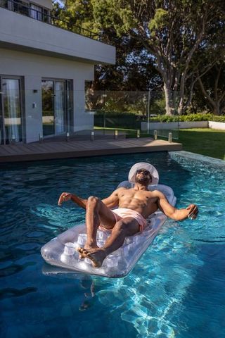Man relaxing on inflatable pool lounger wearing sunhat and sunglasses in backyard pool