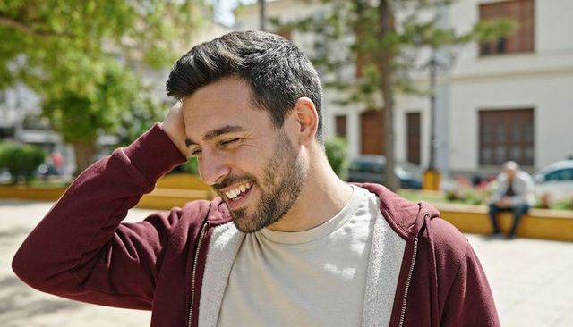 Smiling young man wearing maroon hoodie laughing and looking down in sunlit urban plaza