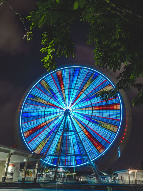 Colorful Ferris Wheel glowing at night against a dark sky in an amusement park. The vibrant lights create a festive and fun atmosphere, making it a perfect image for highlighting amusement parks, tourism promotions, festive events, and vibrant carnival scenes. The image suggests entertainment, thrill, and excitement, ideal for travel brochures, posters, and social media campaigns.