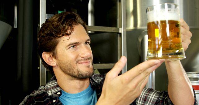 Young man examining a freshly brewed glass of beer in a brewery. Ideal for illustrating concepts related to beer tasting, brewing processes, small-batch breweries, and craft beer industries. Perfect for articles or promotions about the beer brewing industry, craft beer culture, and beer quality assurance.