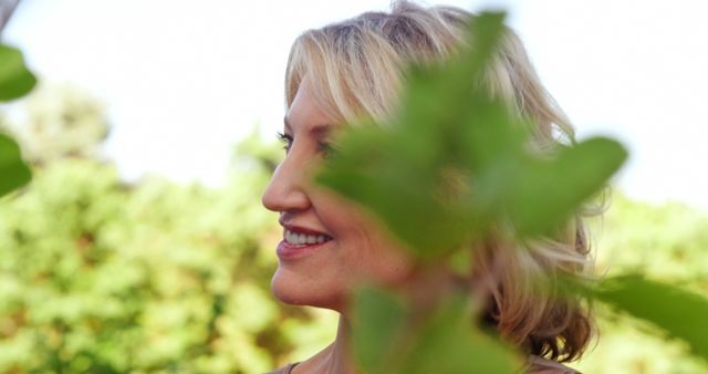 A middle-aged Caucasian woman smiles gently, with copy space to the left amidst a backdrop of green foliage. Her cheerful expression and the natural setting convey a sense of tranquility and happiness.