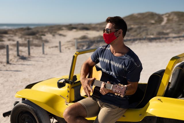 Man wearing face mask sitting on yellow beach buggy playing guitar on a sunny beach. Ideal for themes related to summer holidays, road trips, COVID-19 safety, outdoor activities, music, and relaxation.