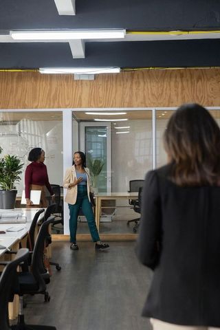 Diverse female coworkers collaborating near glass-walled meeting room in open-plan office