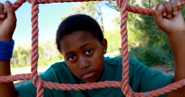 This scene shows a young boy in a green shirt climbing a red rope net in an outdoor park setting on a sunny day. Ideal for illustrating active childhoods, outdoor adventures, and playful moments. Perfect for use in educational materials, promotional content about parks and recreational activities, or articles about child development and fitness.