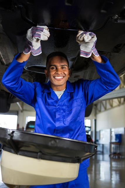 Mechanic in blue uniform smiling while servicing a car in a repair garage. Ideal for use in automotive service advertisements, mechanic training materials, and promotional content for car repair shops.