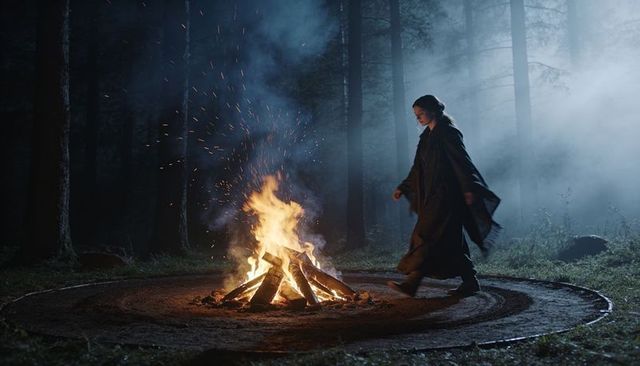 Cloaked woman walking around bonfire in foggy forest clearing at night, sparks rising