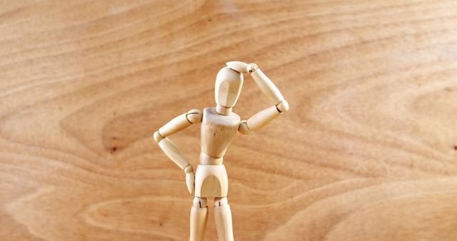 A wooden artist's mannequin poses thoughtfully against a warm wood grain background, with copy space. Its hand-on-head gesture suggests contemplation or confusion, often used to represent creativity or problem-solving.