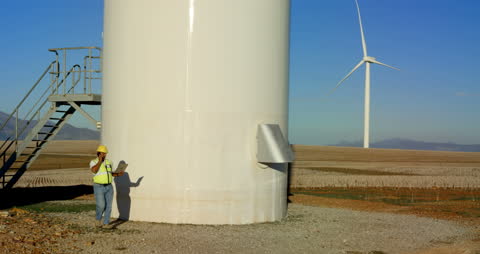 Engineer Inspecting Wind Turbine Tower with Laptop in Open Field