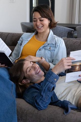 Female friends relaxing on sofa, reading brochures and laughing together at home