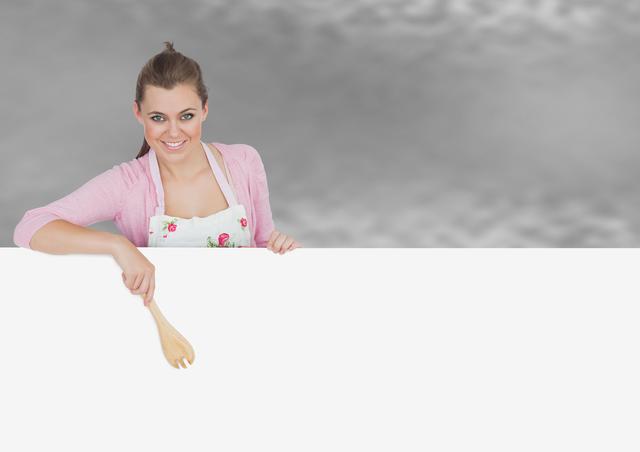 Woman holding a wooden spoon while standing behind a large, blank poster, wearing a light pink sweater and floral apron. Ideal for promotional material, cooking classes, recipe blogs, advertisements, or educational content in culinary arts.